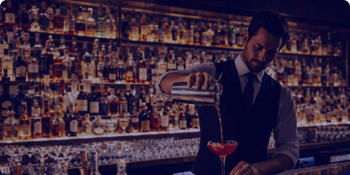 A busy, well-stocked bar at night, with back lit bottle shelves. A bar tender pours a cocktail - FLOX